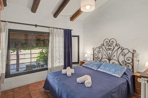 Dormitorio en Can Gat Vell con cabecero de hierro forjado, ropa de cama azul, suelos de barro, vigas vistas y acceso a patio