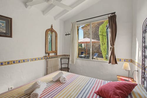 Dormitorio luminoso con ropa de cama de rayas coloridas, techo con vigas de madera y vistas al jardín a través de grandes ventanales en Can Gat Vell