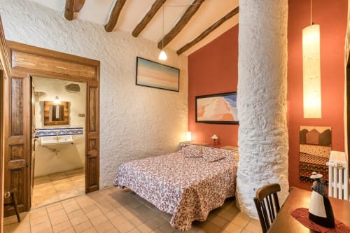 Dormitorio rústico en la casa rural Can Gat Vell en Cataluña con paredes de piedra blanca, vigas de madera, pared de acento naranja y decoración tradicional