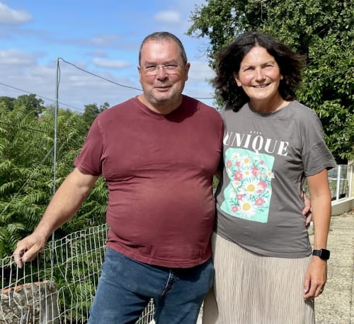 Deux hôtes souriants du Gîte Le Petit Moulin à Pouzauges, France, debout dehors près de la clôture du jardin