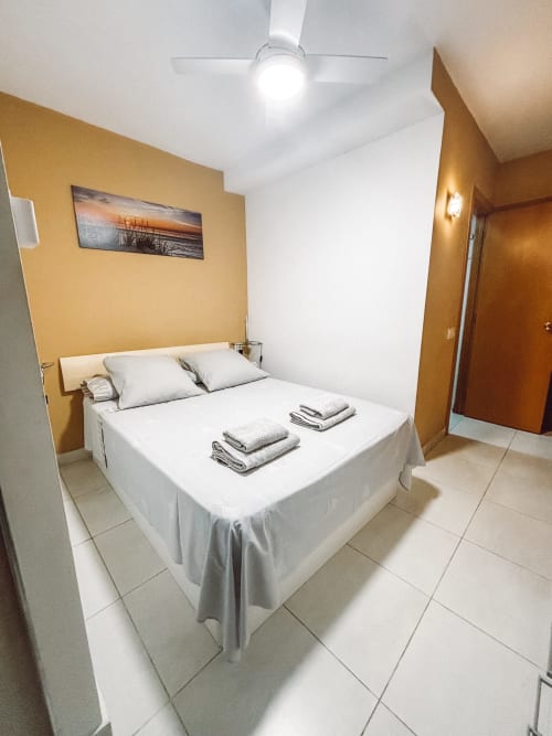 Dormitorio moderno en apartamento La Ventana, Costa Brava, con cama blanca, paredes amarillas, ventilador de techo y cuadro de atardecer