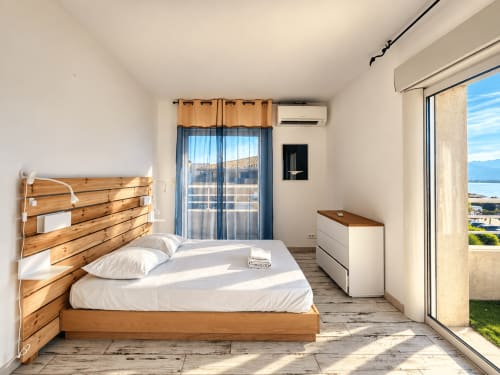 Chambre moderne lumineuse avec lit en bois, draps blancs, vue sur la mer par portes vitrées et terrasse en front de mer