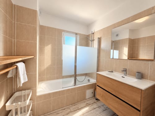 Salle de bain moderne avec carrelage beige, meuble vasque en bois, baignoire avec paroi de douche vitrée et lumière naturelle à L'Île-Rousse