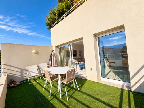 Terrasse moderne d'un appartement face mer à L'Île-Rousse avec vue sur la mer, table à manger blanche, chaises longues et gazon synthétique