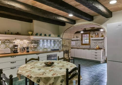 Cocina rústica en casita catalana del siglo XVIII con vigas vistas, azulejos decorados y entrada de arco redondeado.