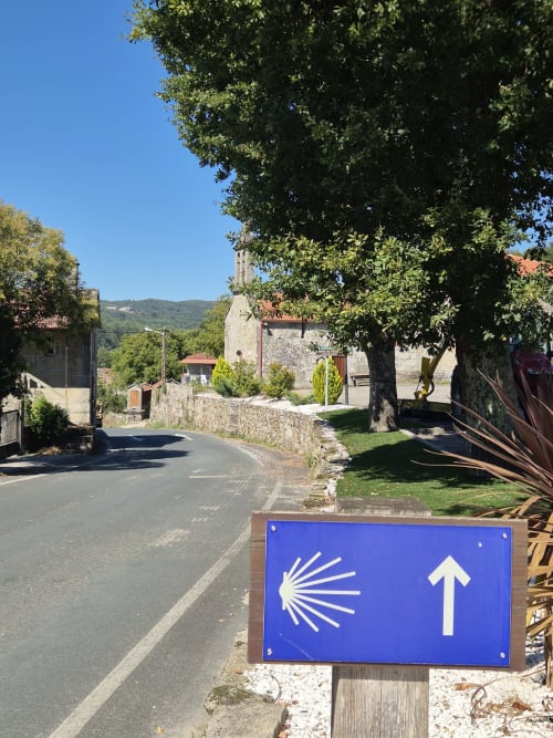 Camino de Santiago waymark sign at rural hotel entrance in Pazos de Arenteiro with stone buildings and mountains