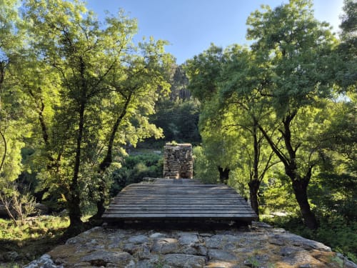 Wooden bridge through lush green trees leading to stone buildings at Aldea Rural Pazos De Arenteiro hotel in Spain
