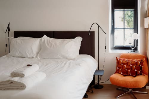 Modern studio bedroom at Castanea Business & Suites with white bedding, dark headboard, orange armchair, and natural window