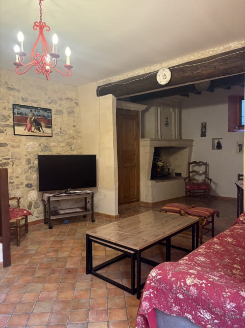 Spacieux salon Arlésien aux murs en pierre, lustre rouge, télévision, cheminée et sièges rouges traditionnels