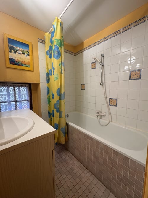 Salle de bains avec carrelage blanc, rideau de douche à fleurs jaunes, lavabo et baignoire dans un gîte à Arles