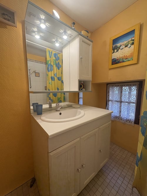 Salle de bains jaune vif dans location à Arles avec meuble-lavabo blanc, miroir éclairé et rideau de douche bleu