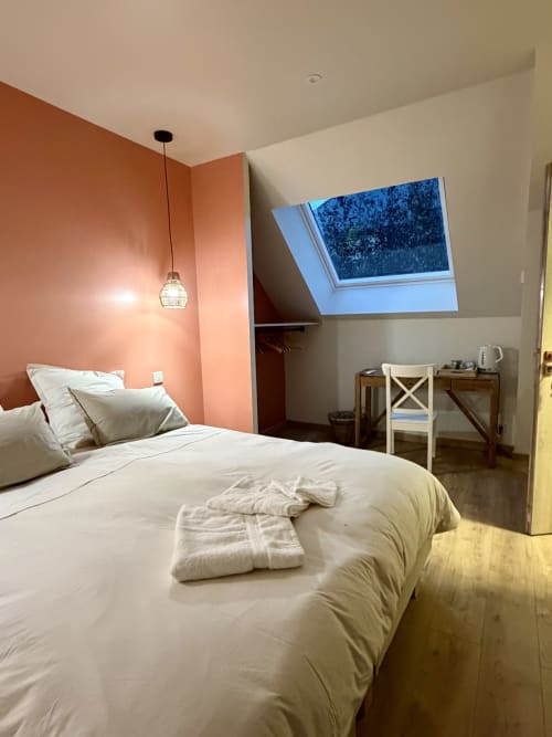 Chambre moderne à La cour des Agapanthes avec mur d'accent corail, puits de lumière, literie blanche, luminaire suspendu et espace de travail.