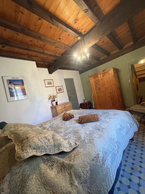 Chambre spacieuse à Arles avec plafond à poutres apparentes, lit garni de courtepointes, meubles en bois et carreaux de céramique bleu motifs