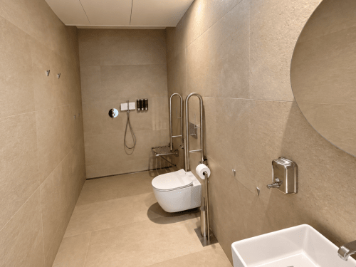 Baño accesible en el hotel Holibai de Baiona con ducha a nivel del suelo, inodoro empotrado en la pared, barras de agarre y alicatado beige