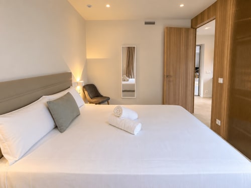 Moderno dormitorio en apartamento Holibai, Baiona con cama blanca, cabecero gris, detalles en madera, espejo e iluminación cálida
