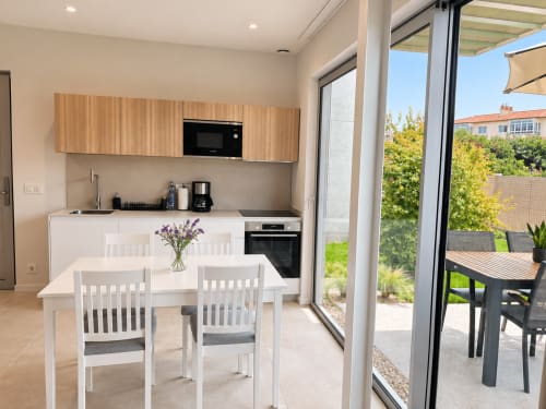 Moderna cocina-comedor en apartamento de Baiona con mesa blanca, muebles de madera y vistas al jardín por puertas correderas