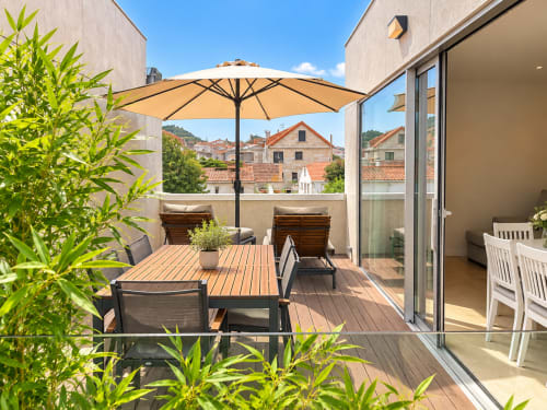 Soleada terraza del apartamento Holibai en Baiona con suelo de madera, mesa de comedor, sombrilla beige, tumbonas y vistas a la ciudad
