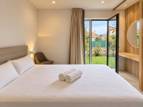 Habitación moderna del hotel Holibai en Baiona con cama de matrimonio, vistas al jardín a través de puertas de cristal, detalles en madera y toallas enrolladas
