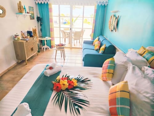 Chambre de l'appartement Ti Oasis Trinidad aux murs turquoise, vue sur mer, coussins colorés, fleurs tropicales et équipements modernes