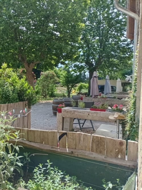 Outdoor patio at Domaine Le Piboul BnB with wooden tables, pink flowers, umbrellas, and lush green gardens in Fontcouverte