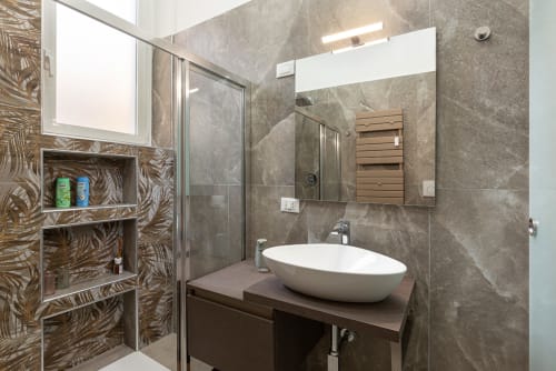 Bagno moderno presso LM Apartments Pavia con lavabo bianco, piastrelle grigie, box doccia e dettagli in legno