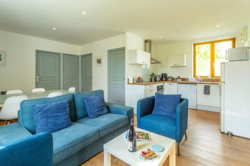 Séjour ouvert avec canapé bleu, chaises de salle à manger, cuisine blanche et vue sur le jardin à l'appartement Clubhouse Gîtes à Beussent,
