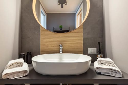 Bagno moderno presso LM Apartments Pavia con lavabo bianco ovale, specchio LED, dettagli in legno e asciugamani ordinati