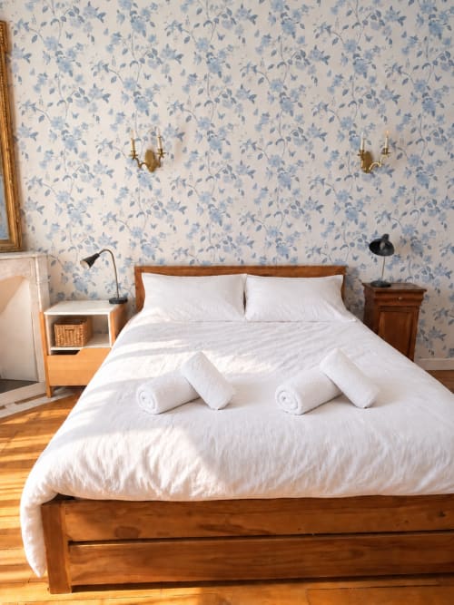 Chambre lumineuse de la villa ARLOÉ à La Rochelle avec lit en bois, draps blancs, papier peint fleuri bleu et tables de nuit en bois.
