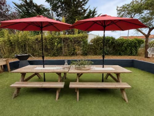 Espace de restauration en plein air à la Villa Alba ARLOÉ avec tables de pique-nique en bois, parasols rouges et vue sur le jardin à La Rochelle