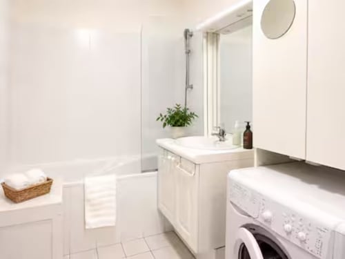 Salle de bain moderne avec vanité blanche, lave-linge, miroir rond et plante en pot à l'appartement ARLOÉ à La Rochelle