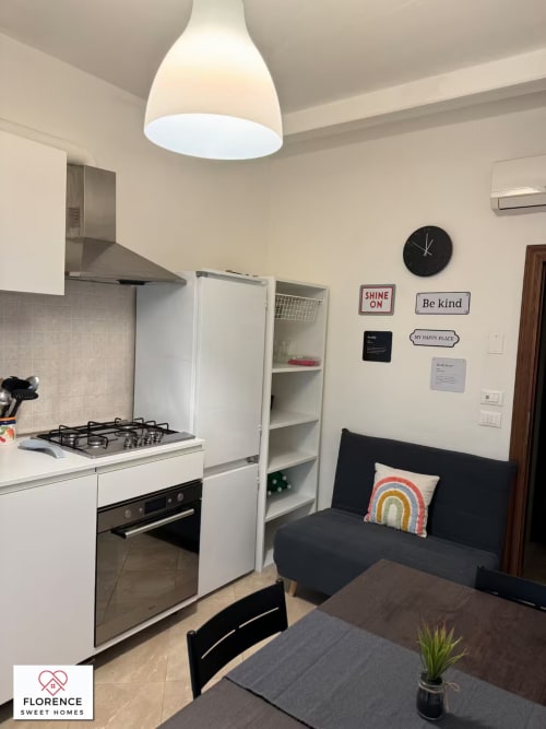 Zona cucina-soggiorno moderna nell'appartamento Florence Sweet Homes con fornello a gas, mobili bianchi, divano nero e lampada a sospensione