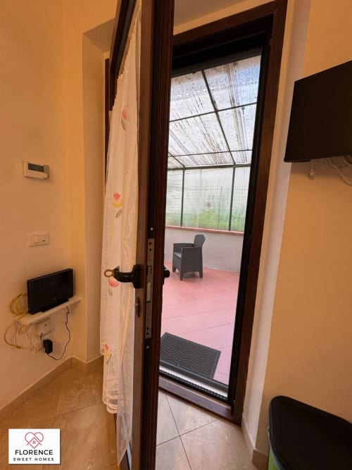Porta in vetro aperta verso la terrazza di un appartamento a Firenze con piastrelle rosa, sedia e vista sulla serra