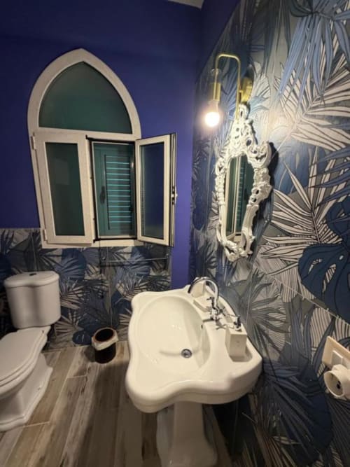 Bagno moderno di Villa Brikinnia con lavabo bianco curvo, toilette, specchio ornamentale e carta da parati tropicale blu a Lentini
