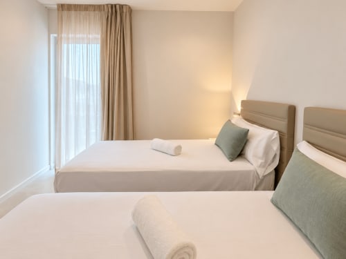 Habitación doble moderna en apartamento de Baiona con cabeceros beige, ropa de cama blanca, almohadas verdes y cortinas de piso a techo
