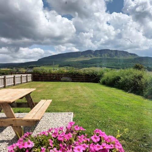 Ballymultimber Cottages garden view with picnic table, pink flowers, green lawn, and mountain backdrop in Limavady