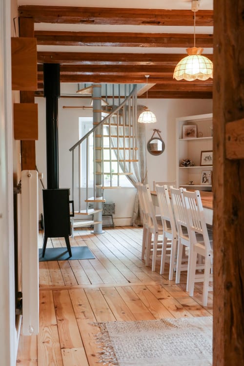 Salle à manger rustique avec poutres en bois, chaises blanches, poêle noir et luminaires suspendus à La Maison du Gardien