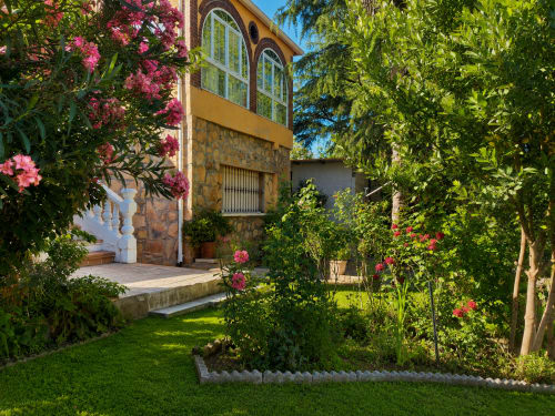 Casa de piedra rústica con contraventanas amarillas rodeada de flores rosadas en flor y vegetación exuberante en los jardines de Madrid
