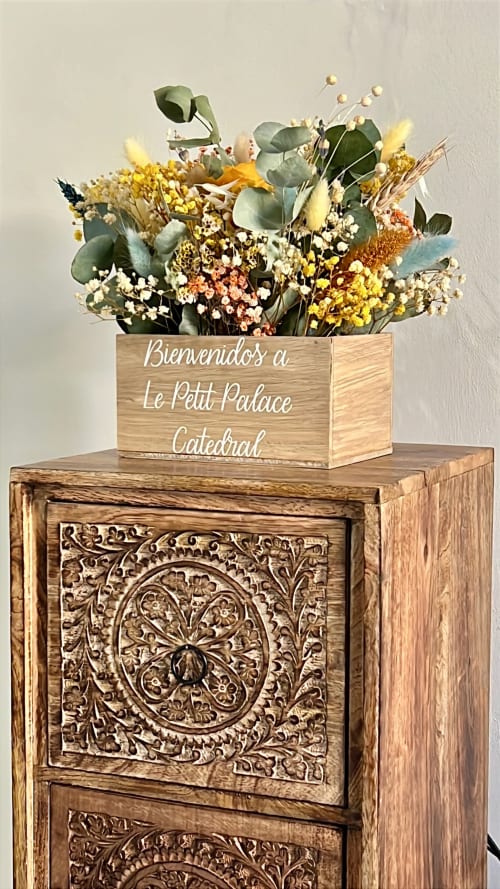 Dried flower arrangement in wooden box reading 'Bienvenidos a Le Petit Palace Catedral' on ornate wooden pedestal