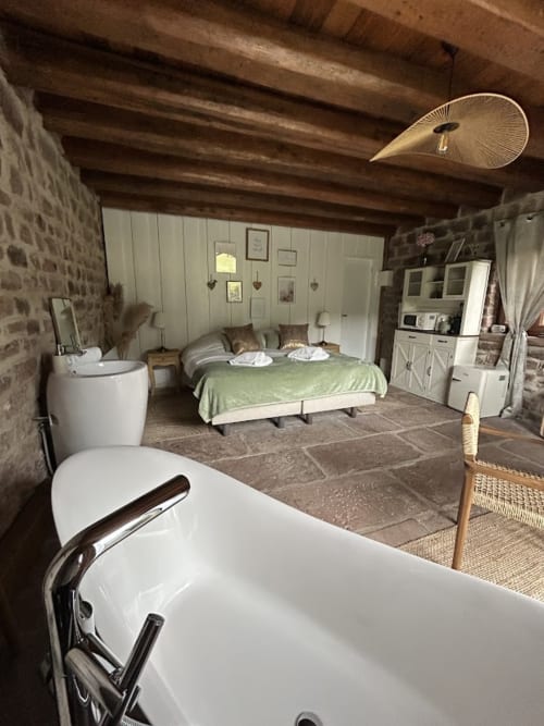 Spacieuse chambre de La Cabane de Grès avec baignoire îlot, linge de lit vert, poutres apparentes et murs en pierre