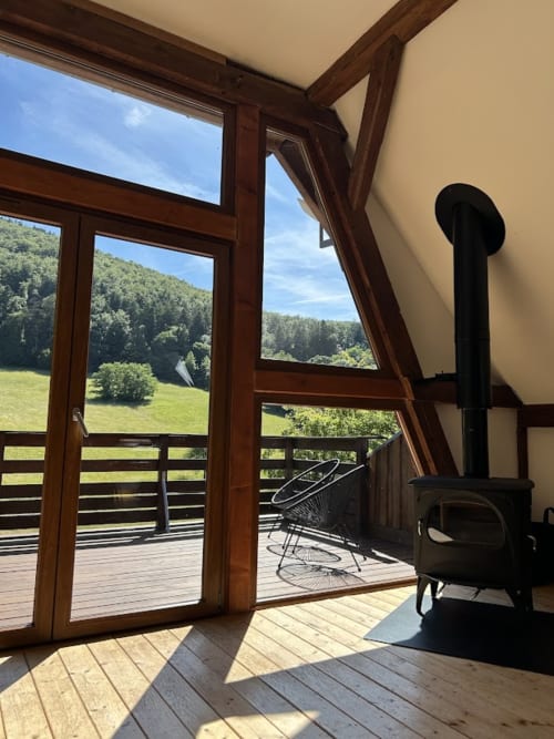 Intérieur lumineux d'un chalet avec portes en bois ouvrant sur une terrasse surplombant une vallée verdoyante et des montagnes boisées aux Cabanes du