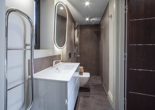 Modern bathroom at The Coach House villa with white sink, oval lit mirror, walk-in shower, and dark tile flooring
