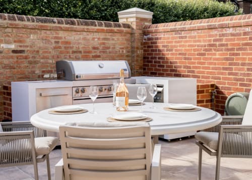 Elegant outdoor dining setup at The Coach House villa with round table, place settings, and built-in kitchen behind brick