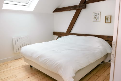 Chambre douillette avec poutres bois apparentes, couette blanche sur lit crème, puits de lumière et décor botanique aux Cabanes du
