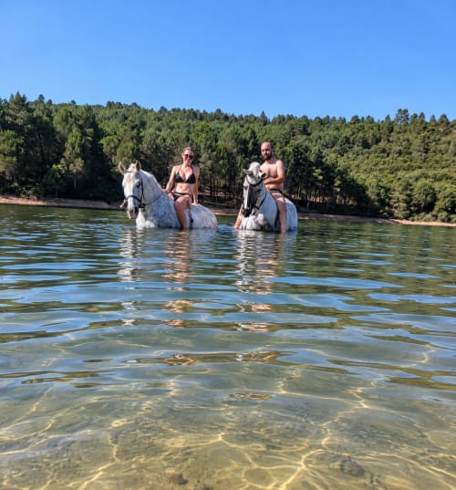 Dos personas cabalgando a caballo blanco en un lago con bosque de fondo en El descanso del peregrino, Guadalupe