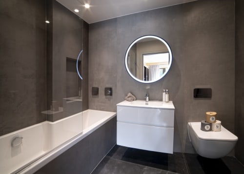 Modern bathroom at The Coach House Hotel featuring circular LED mirror, white vanity, walk-in shower, and dark tile walls