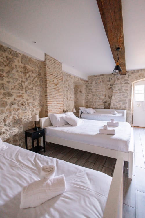 Chambre rustique en pierre avec trois lits, draps blancs, poutres en bois apparent et serviettes roulées aux Villas Bacchus