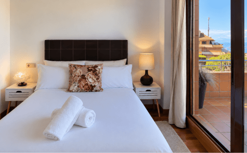 Dormitorio luminoso en apartamento Holibai con vistas al mar, ropa de cama blanca, cabecero oscuro y acceso a terraza en Baiona
