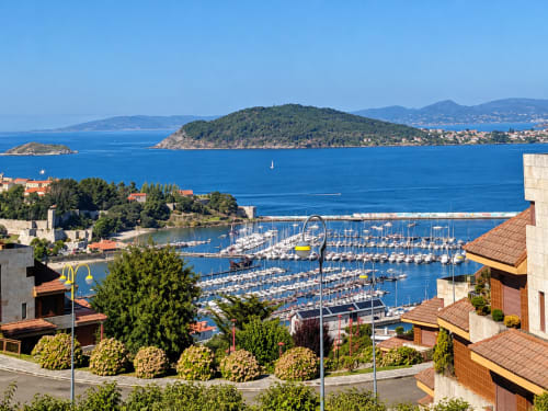 Marina llena de veleros en Bañoa, España con pueblo costero, isla boscosa verde y mar azul cristalino bajo cielos soleados