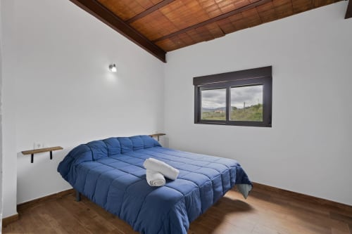 Dormitorio amplio en la casa rural Holiday Dreams Ronda con edredón azul, techo de madera y vistas a las montañas por la ventana