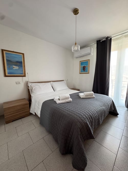 Modern bedroom in Sormano apartment with queen bed, white linens, gray quilted coverlet, pendant light, and framed coastal